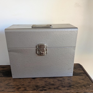 May include: A rectangular, silver-colored metal storage case with a textured finish. The case features a hinged lid, a silver metal carrying handle on the top, and a central silver metal latch mechanism on the front for securing the closure.