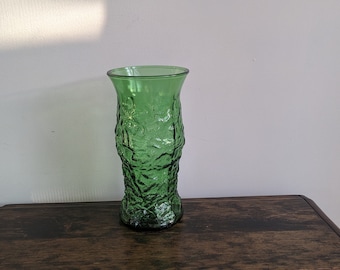 Puede incluir: Un jarrón de vidrio verde con un diseño texturizado similar a la corteza. El jarrón tiene una abertura ancha y una base verde oscuro. Se exhibe sobre una superficie de madera oscura contra una pared blanca.