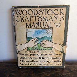 May include: Vintage "Woodstock Craftsman's Manual" book. The cover features the title in large, black, distressed lettering. A central illustration depicts hands crafting, with a landscape backdrop. The book's subtitle lists various crafts, including weaving and pottery.