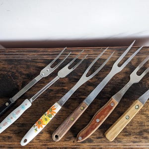 Può includere: Una collezione di forchette da intaglio vintage con denti in acciaio inossidabile e vari design del manico. I manici includono stili neri, floreali e in legno. Le forchette sono disposte su una superficie di legno scuro.