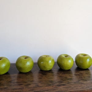 May include: Five smooth, round, artificial apples in a vibrant shade of lime green are arranged in a horizontal row. Each apple features a subtle, realistic texture with slight color variations and a small, brown, recessed stem at the top center.