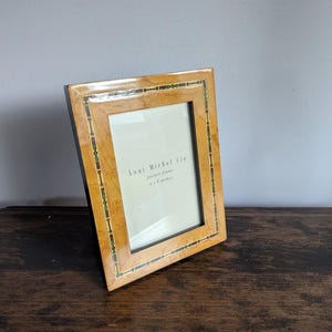 Può includere: Una cornice rettangolare con finitura a venatura di legno chiaro e dettagli decorativi verdi e dorati. La cornice è etichettata "Loui Michel Cie" ed è progettata per una foto di 10 x 15 cm. La cornice è esposta su una superficie di legno scuro.