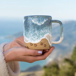 May include: A ceramic mug with a beach design. The top half is blue and white, mimicking ocean waves, and the bottom half is tan, like sand. The mug has a handle and is decorated with horseshoe imprints.