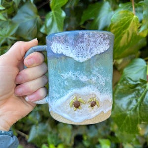 Puede incluir: Una taza de cerámica hecha a mano con un diseño costero. La taza presenta un patrón de olas azules y blancas, con dos pequeñas figuras de tortugas en una playa de arena. La taza se sostiene contra un fondo de follaje verde.