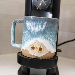 May include: A ceramic mug featuring a textured blue and white glaze that mimics ocean waves crashing onto a sandy beige shore. Two small, sculpted yellow sea turtles with brown shells are attached to the lower portion of the mug's exterior.