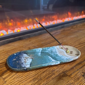 May include: Handmade ceramic incense holder with a blue and white wave design. The oval-shaped dish has a light brown section with paw print details. An incense stick is lit, emitting smoke, and a small yellow bead is attached.