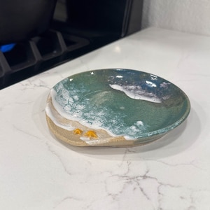 Może przedstawiać: Ceramiczny podkładka pod łyżkę w kształcie plaży z zielonym oceanem, białymi falami i żółtymi muszelkami.