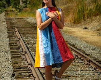 May include: A woman wearing a colourful striped dress stands on a set of railway tracks. She is smiling and holding her hands to her chest.