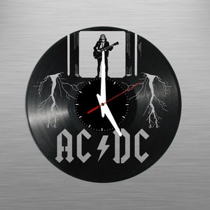 Puede incluir: Reloj de vinilo negro con una silueta blanca de un músico tocando la guitarra frente a un edificio con pilares. La esfera del reloj es negra con agujas blancas y las palabras "AC/DC" están impresas en blanco debajo de la esfera.