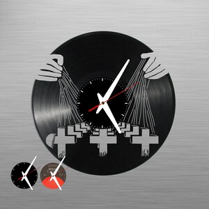 May include: A black vinyl record clock with a white clock face and hands. The record is decorated with a black and white image of a band's album cover. The album cover features a group of crosses and a pair of hands reaching up.