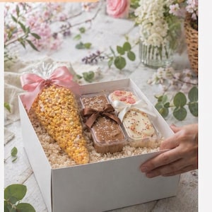 May include: A white gift box filled with fudge and a large bag of caramel corn, tied with a pink ribbon. The box contains several fudge pieces in clear containers. Text on the box reads "Gourmet Fudge Gift Box".