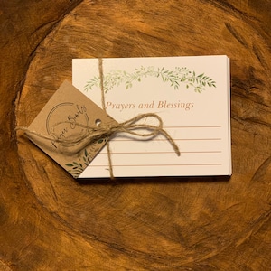 May include: A stack of white cards with a green floral border and the text "Prayers and Blessings" on the top card. The cards are tied together with twine and a brown paper tag with the text "Prayers & Bulbs" on it.