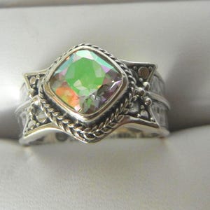 May include: A silver ring with a square, iridescent gemstone. The gemstone is framed by a twisted rope design and set in a detailed, textured band. The ring's design includes small, decorative elements.
