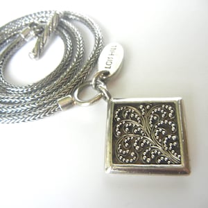May include: Silver-toned necklace with a square pendant featuring an ornate, swirling filigree design. The pendant is framed with a silver border. The chain is a woven style, and the necklace is displayed on a white surface.