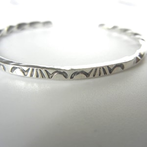 May include: A silver cuff bracelet with a textured, hammered finish. The bracelet features a twisted rope design on the ends and a repeating pattern of curved lines and triangles along the front. The jewelry is displayed on a white surface.