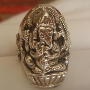 Może przedstawiać: Szczegółowy srebrny pierścień przedstawiający rzeźbę hinduskiego bóstwa Ganesha. Pierścień prezentuje misterne detale boga z głową słonia w pozycji siedzącej, z teksturowanym wykończeniem i szeroką obrączką.