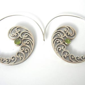 May include: A pair of silver spiral earrings with a swirling filigree design. Each earring features a small, round, light green gemstone. The earrings are set against a white background.