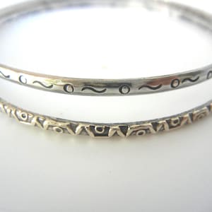 Puede incluir: Dos pulseras de plata. La pulsera superior tiene una serie de diseños ondulados y circulares grabados. La pulsera inferior tiene un patrón geométrico repetitivo. Las joyas se exhiben sobre una superficie blanca.