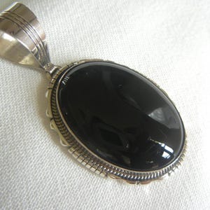 May include: Oval black onyx pendant with a silver-tone bail. The pendant has a polished, glossy surface and is set in a decorative silver frame. The bail is also silver and features etched lines. The pendant is displayed on a white surface.