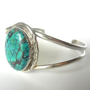 May include: A silver cuff bracelet featuring a large, oval turquoise stone with dark matrix patterns. The stone is set in a decorative bezel, and the bracelet has a simple, open design.