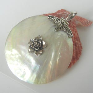 May include: A large, circular, iridescent mother-of-pearl pendant featuring a central silver flower embellishment and an ornate silver filigree bail at the top. The pendant is layered over a textured, fan-shaped, reddish-orange seashell.