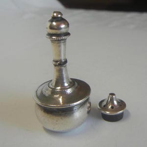 Può includere: Un piccolo contenitore in metallo color argento con un collo lungo e sottile e una base arrotondata. Il contenitore ha un coperchio rimovibile con piccoli fori. Il design è ornato e dettagliato, suggerendo uno scopo decorativo o cerimoniale.
