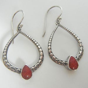 May include: A pair of silver teardrop earrings with a red coral stone at the bottom. The earrings have a detailed, textured design with small beaded accents around the edges and decorative swirls.