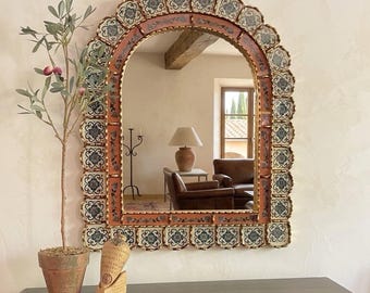 Op de afbeelding: Een boogvormige spiegel met een decoratief frame met een herhalend bloemen tegelpatroon in blauw, wit en goud. Het frame heeft een terracotta-kleurige achtergrond. Een kleine potplant en een decoratief beeldhouwwerk staan op een donkere houten ondergrond.