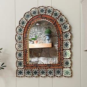 May include: An arched mirror with a decorative frame. The frame features a terracotta-colored border with floral tile accents in white, teal, and gold. The mirror reflects a stone wall and a shelf with plants.