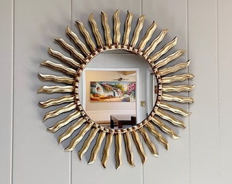 May include: A gold sunburst mirror with a round mirror in the center. The mirror reflects a room with a painting on the wall.