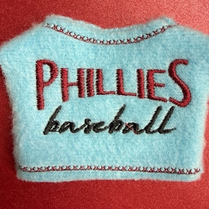 May include: A light blue felt baseball shirt with the word "PHILLIES" embroidered in red and "baseball" in black. The shirt has a red stitched border and is set against a red background.