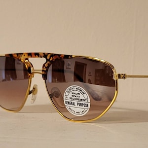 Peut inclure: Lunettes de soleil aviateur dorées avec des verres dégradés bruns et une monture en écaille de tortue. Les lunettes de soleil ont un autocollant sur la lentille droite qui indique "Blocks Minimum 99.9% UV Rays, Meets ANSI Z80.3 Requirements, General Purpose".