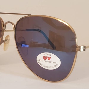 Puede incluir: Gafas de sol aviador doradas con lentes marrones y una pegatina que dice "OPTIMUM UV EYE PROTECTION AGAINST ULTRA-VIOLET RAYS".