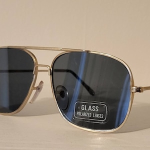 May include: Gold-coloured aviator sunglasses with dark blue polarised lenses. The sunglasses have a double bridge and a rectangular shape. The lenses have a reflective quality. The text on the lens reads "GLASS POLARIZED LENSES".