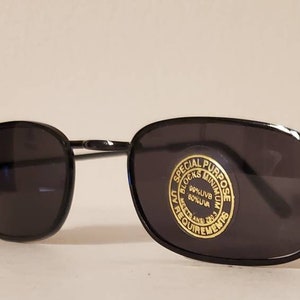Op de afbeelding: Zwarte rechthoekige zonnebril met een zwart metalen montuur. De rechterlens heeft een gouden sticker met de tekst "Special Purpose Blocks Minimum Harmful Rays UV 400", met een cirkel rond de tekst.
