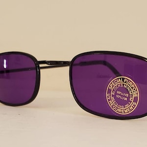 May include: Black-framed sunglasses with dark purple lenses. A circular sticker on the right lens reads: SPECIAL PURPOSE BLOCKS MINIMUM 99% UVB 50% UVA MEETS ANSI Z80.3 UV REQUIREMENTS. The glasses feature thin metal temples and a classic oval shape.