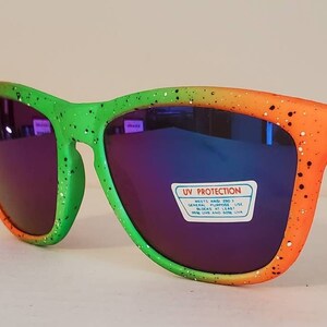 Puede incluir: Gafas de sol con motas verdes, naranjas y amarillas con lentes azules espejados. Las gafas de sol tienen una etiqueta de protección UV que dice "Protección UV, Cumple con ANSI Z80.3, Uso general, Bloquea el 99% o más de los rayos UVA, UVB y UVC".