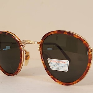 May include: Round sunglasses featuring a tortoiseshell pattern frame with gold-tone metal accents and dark green lenses. A rectangular sticker on the right lens reads UV PROTECTION and includes text regarding ANSI Z80.3 general purpose requirements.