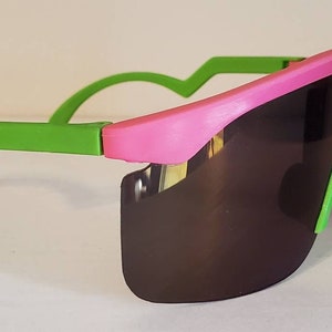May include: A pair of pink and green sunglasses with a wraparound design. The lenses are dark brown and the frame is made of plastic.