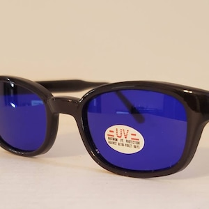 Op de afbeelding: Zwarte plastic zonnebril met blauwe glazen. De zonnebril heeft een label met de tekst "=UV= WARNING EYE PROTECTION AGAINST ULTRA VIOLET RAYS"