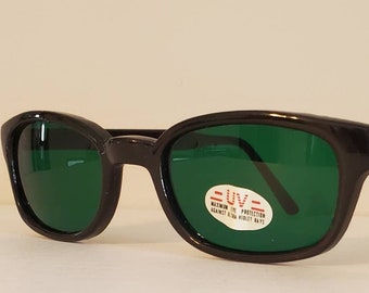 Könnte beinhalten: Schwarze Plastik-Sonnenbrille mit grünen Gläsern. Die Sonnenbrille hat ein Etikett mit der Aufschrift "UV - Maximaler UV-Schutz gegen ultraviolette Strahlen".