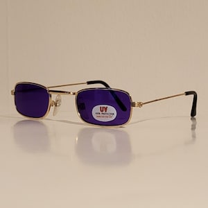 May include: Gold-framed sunglasses with rectangular purple lenses. The sunglasses have a small UV protection sticker on the right lens. The arms are thin and gold, with black tips.