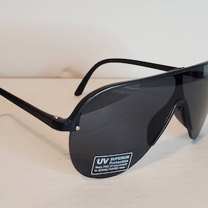 Op de afbeelding: Zwarte zonnebril met een wrap-around ontwerp. De lenzen zijn donkergrijs en de monturen zwart. De zonnebril heeft een sticker met "UV Superior Protection".