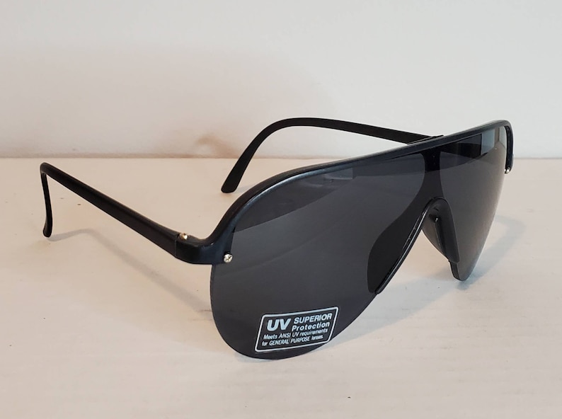 Op de afbeelding: Zwarte zonnebril met een wrap-around ontwerp. De lenzen zijn donkergrijs en de monturen zwart. De zonnebril heeft een sticker met "UV Superior Protection".