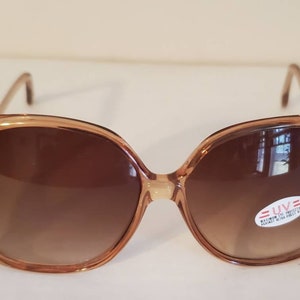May include: Oversized brown sunglasses with a gradient lens. The frames are a light brown color with a slight cat-eye shape. A small sticker on the lens indicates UV protection. A stylish accessory for eye protection.