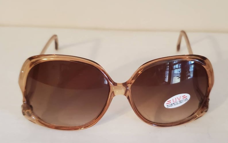 May include: Oversized brown sunglasses with a gradient lens. The frames are a light brown color with a slight cat-eye shape. A small sticker on the lens indicates UV protection. A stylish accessory for eye protection.