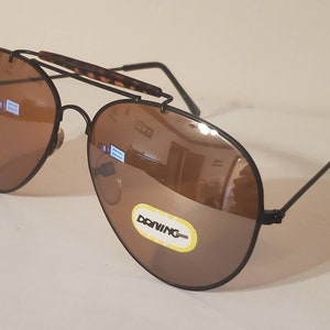 May include: Black aviator sunglasses with brown lenses. The sunglasses have a metal frame and a tortoise shell nose bridge. The sunglasses are labeled "DONNING" on the right lens.