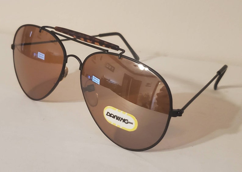 May include: Black aviator sunglasses with brown lenses. The sunglasses have a metal frame and a tortoise shell nose bridge. The sunglasses are labeled "DONNING" on the right lens.