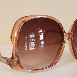 May include: Oversized, light brown sunglasses with gradient brown lenses. The frames are a translucent, light brown color. The lenses reflect the surrounding environment. A small sticker is on the right lens.