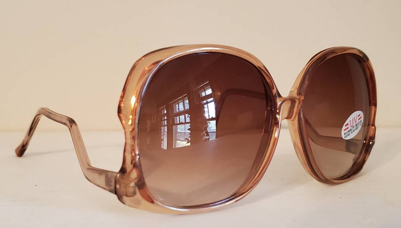 May include: Oversized, light brown sunglasses with gradient brown lenses. The frames are a translucent, light brown color. The lenses reflect the surrounding environment. A small sticker is on the right lens.